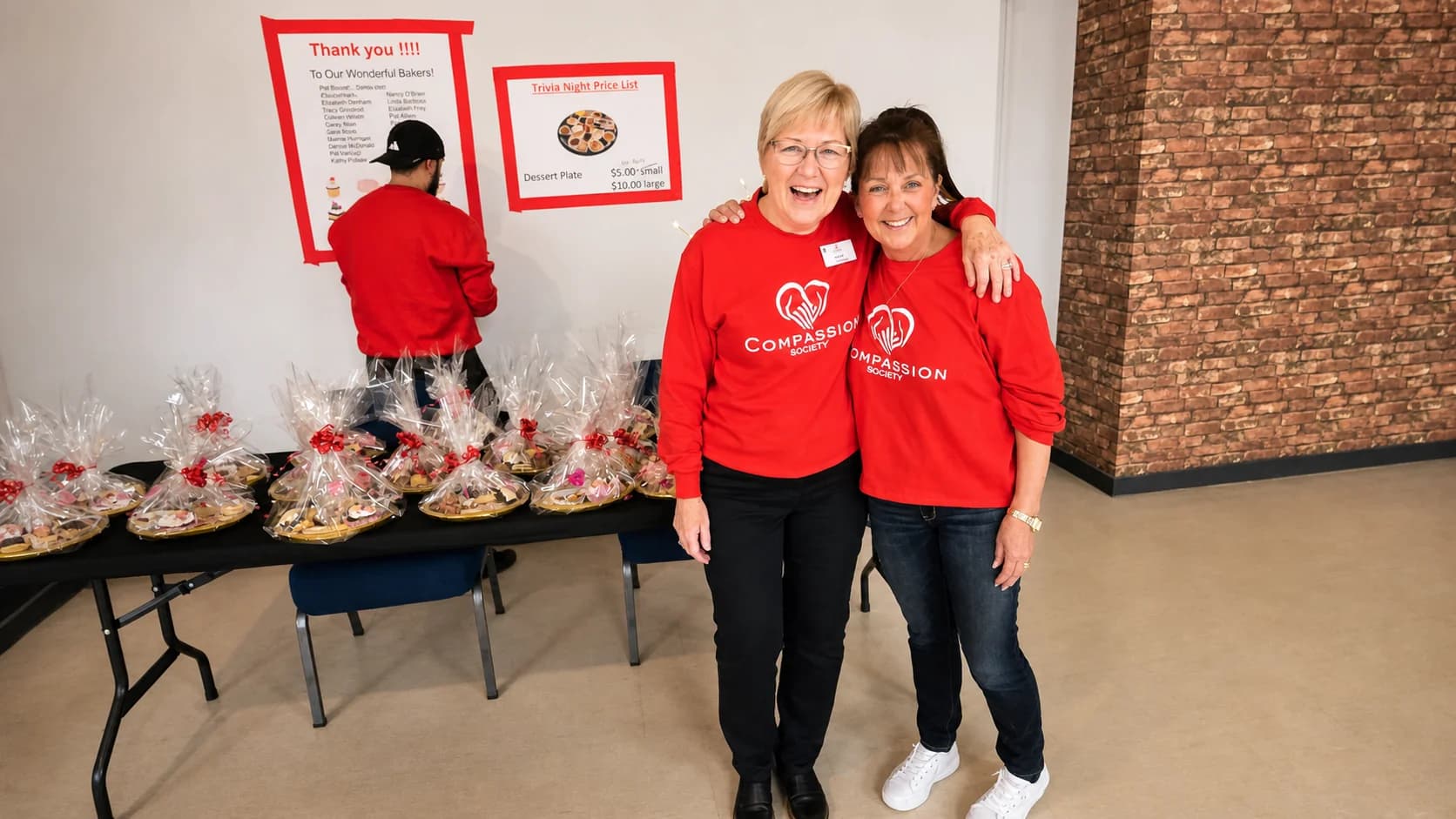 Two Compassion Society of Halton volunteers stand together at a real community fundraiser while a third volunteer works beside a table of prepared baked goods.