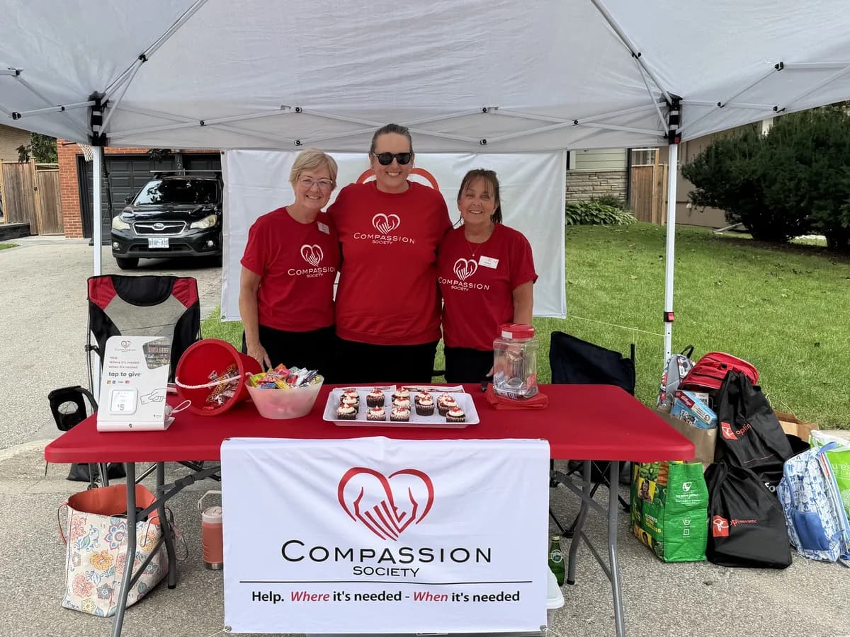 Three Compassion Society of Halton volunteers in red Compassion shirts smile behind a community table under a white tent.