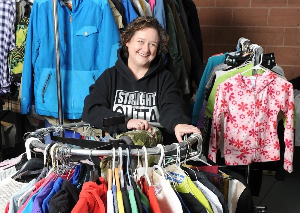 Mina Wahidi standing behind a rack of donated clothing, smiling, surrounded by hanging jackets and children's clothing.