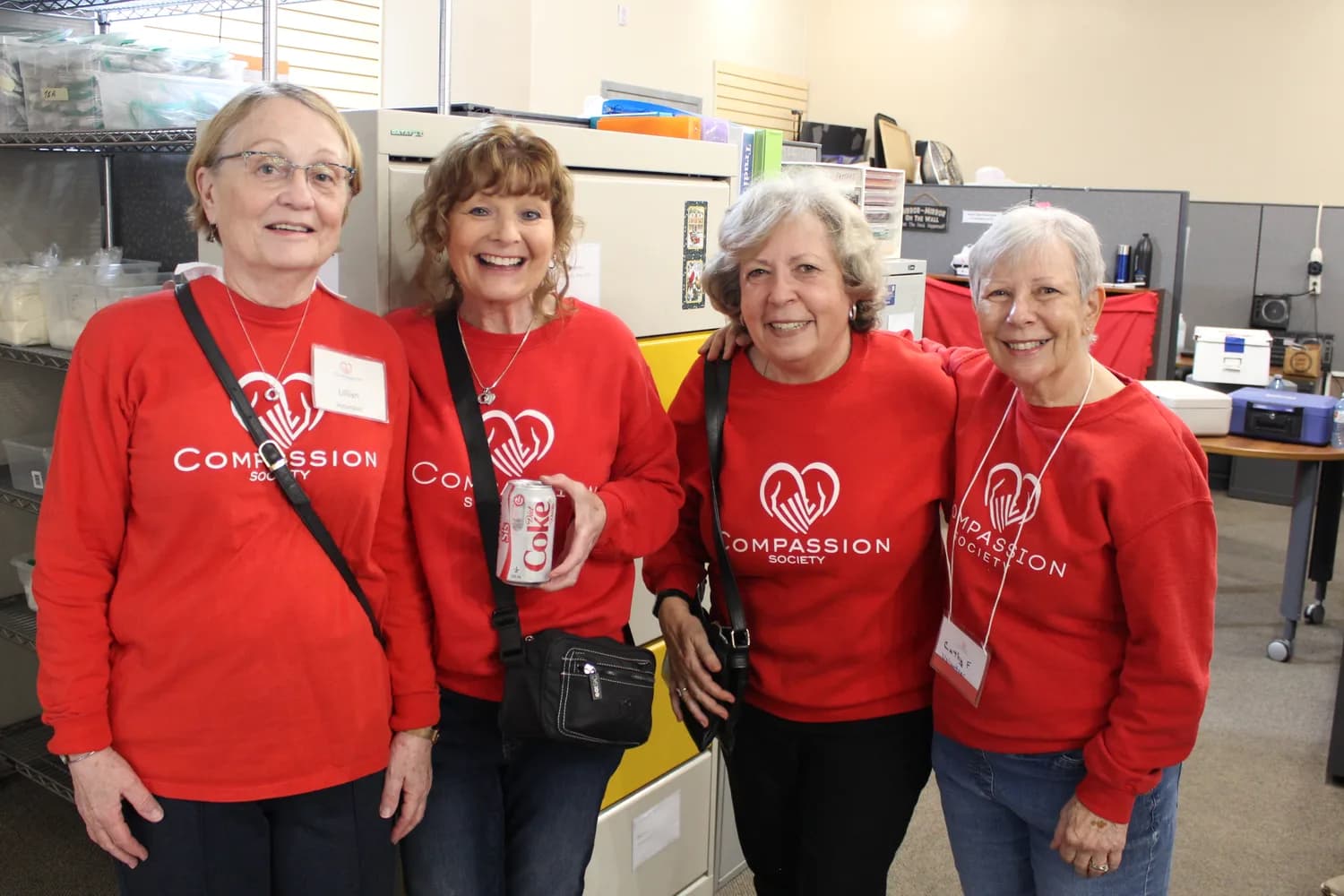 Four Compassion Society of Halton volunteers stand together in matching red Compassion Society sweatshirts in the donation office, two identified by name tags reading Lillian, Volunteer and Cathy F., Volunteer.