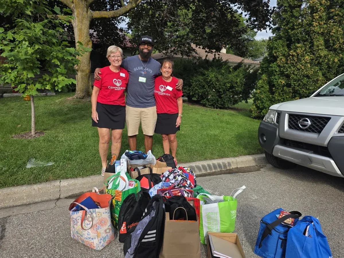 Two Compassion Society of Halton volunteers and a drive organizer stand behind bags of donated food and essentials.