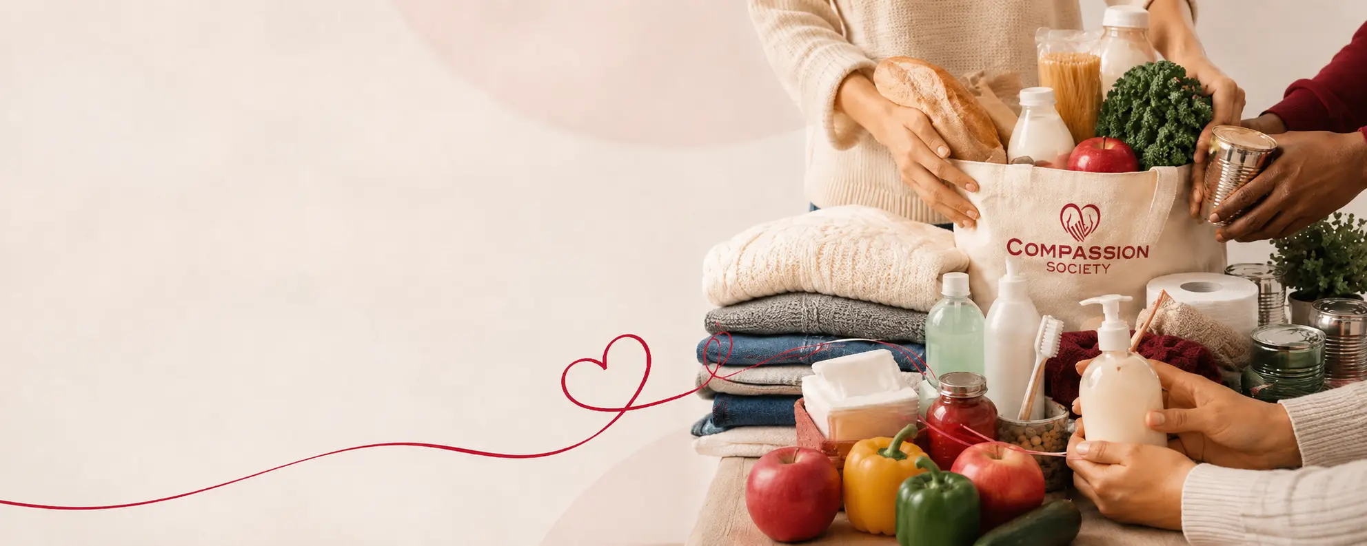 Hands packing bread, milk, fresh produce and pantry staples into a canvas Compassion Society tote bag, with folded clothing, personal care items and household essentials laid out nearby.