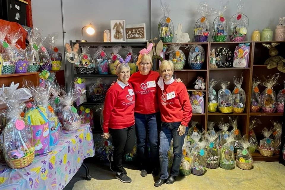 Three Compassion Society of Halton volunteers wearing matching red branded sweatshirts stand together in a stockroom filled with wrapped Easter baskets ready for distribution.