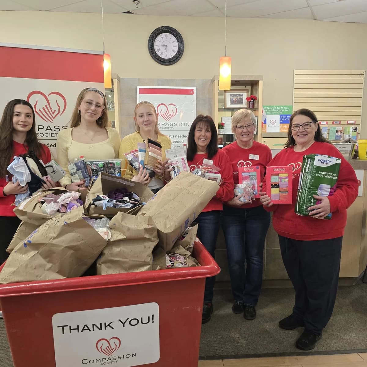 Six Compassion Society of Halton volunteers and staff in red and yellow CSH sweaters, smiling behind a donation bin filled with donated hygiene and clothing items, marked with a Thank You sign.