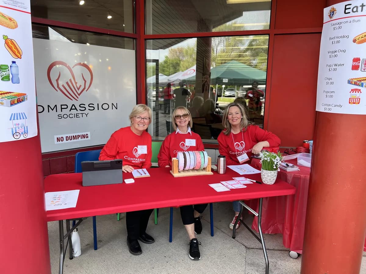 Three Compassion Society of Halton volunteers in red Compassion sweatshirts welcome people at a community fundraiser table.
