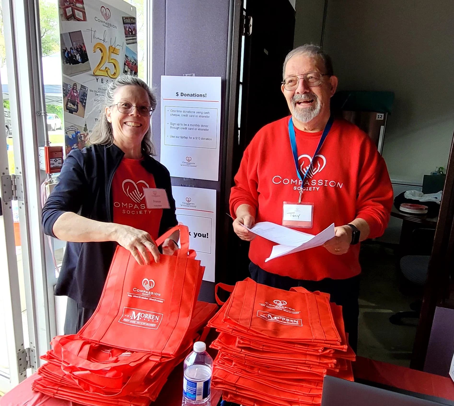 Two Compassion Society of Halton volunteers stand together at a table stacked with branded reusable tote bags, beside a sign describing ways to give a monetary donation.