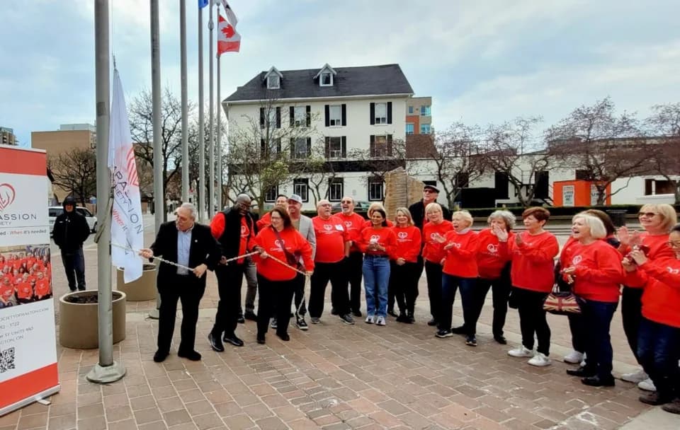 A large group of Compassion Society of Halton volunteers in red Compassion sweatshirts gather outdoors for a community flag raising.