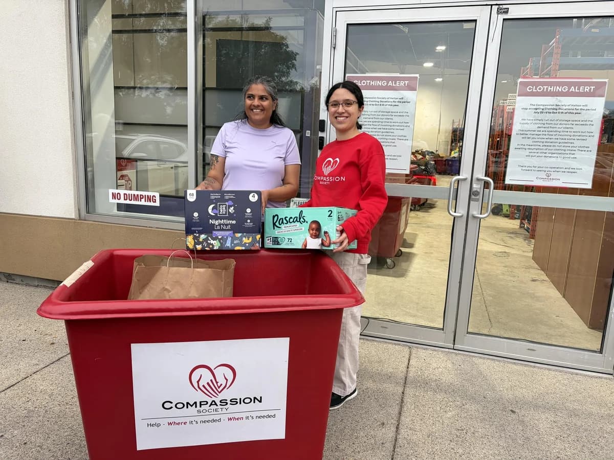 A donor and a Compassion Society of Halton volunteer hold up packs of diapers beside a red Compassion donation bin at the warehouse door.