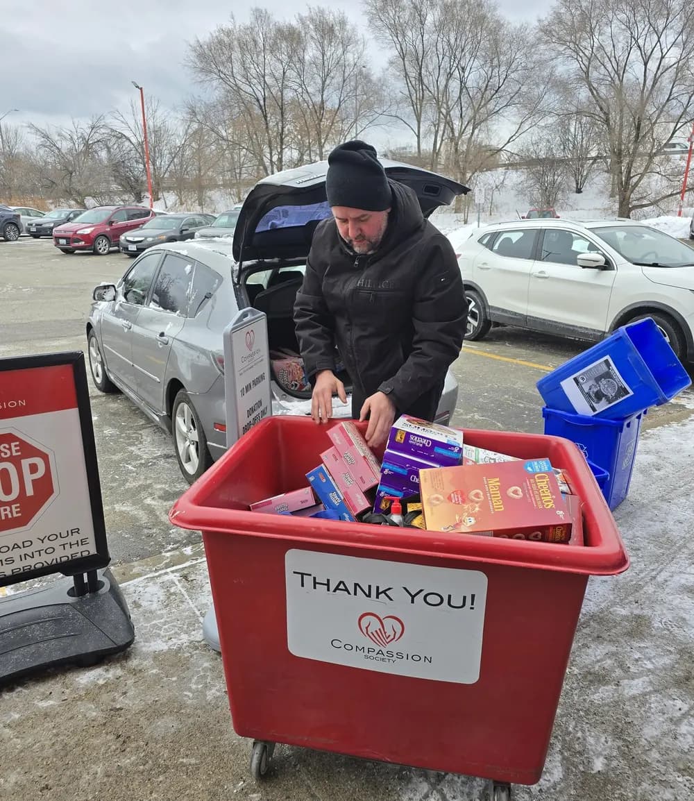 A donor places grocery boxes into a red Compassion Society of Halton donation bin marked Thank You, parked beside their car in the Donation Warehouse lot on a winter day.